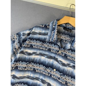 Puede incluir: Una camisa de manga corta con botones en tonos azul, blanco y negro. La tela presenta un patrón repetitivo de la palabra SOUTHPOLE en texto blanco, intercalado con bandas horizontales de cuadrados pixelados y líneas verticales degradadas.