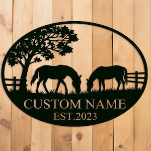 Puede incluir: Letrero ovalado de metal negro con dos caballos pastando en un campo, con un árbol y una valla. El letrero incluye el texto "CUSTOM NAME" y "EST.2023". El fondo es una superficie de madera.