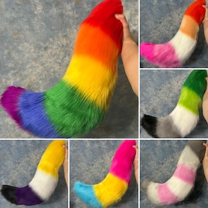May include: A collection of six faux fur tails in various colour combinations. The tails feature layered, multi-coloured designs including a rainbow gradient, a pink-white-orange gradient, a green-white-black pattern, and other tri-colour arrangements.