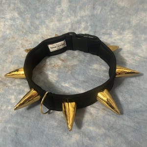 May include: A black collar featuring large, shiny gold-toned spikes arranged around the band. The collar includes a black plastic side-release buckle and a gold metal D-ring. A white fabric label inside reads HANDMADE BY KAHANI CREATIONS 2026.