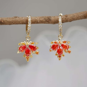 May include: A pair of gold hoop earrings featuring dangling maple leaf charms. Each leaf is encrusted with vibrant orange and red teardrop-shaped stones, accented by smaller yellow and orange crystals. The hoops are lined with clear white stones.