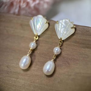May include: A pair of gold earrings with white shell and pearl accents. The shell is shaped like a fan and the pearls are round. The earrings are hanging from a gold chain.