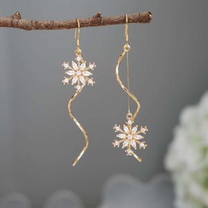 May include: A pair of gold-coloured earrings. Each earring features a snowflake design with clear, sparkling stones. The earrings hang from a curved, gold-toned wire, creating a delicate and elegant look. The earrings are displayed against a soft grey background.