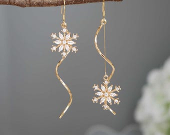 May include: A pair of gold-coloured earrings. Each earring features a snowflake design with clear, sparkling stones. The earrings hang from a curved, gold-toned wire, creating a delicate and elegant look. The earrings are displayed against a soft grey background.