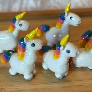 May include: A collection of six small, white unicorn figurines with yellow horns and rainbow manes and tails. The unicorns are arranged on a wooden surface, with a light-colored background.