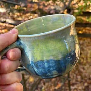 May include: A handmade ceramic mug with a unique glaze. The mug features a blend of green, blue, and cream colors. The mug has a rounded shape and a sturdy handle, perfect for enjoying your favorite beverage.