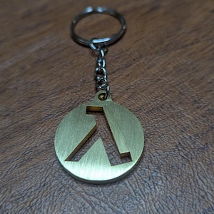 Puede incluir: Un llavero de metal dorado con un colgante circular con un diseño geométrico estilizado. El colgante está unido a una cadena y a un llavero plateado. El fondo es una superficie marrón texturizada.