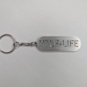 以下が含まれることがあります： キーリングと「HALF-LIFE」という文字がくり抜かれた長方形のタグが付いた、銀色の金属製のキーホルダーです。