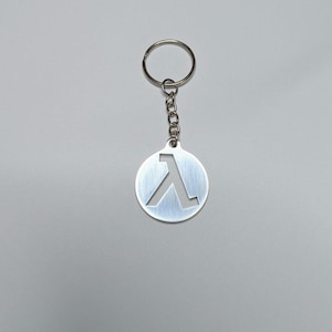 Puede incluir: Un llavero plateado con un colgante circular con un símbolo lambda recortado. El colgante está unido a una cadena y un llavero. El fondo es de color gris claro liso.