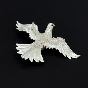 May include: A silver-colored dove brooch with outstretched wings, adorned with small, clear crystals. The bird is set against a black background, showcasing its detailed feather design and overall elegant appearance.