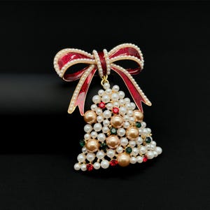 May include: A Christmas tree brooch with a red bow and pearl accents. The tree is made of white, gold, red, and green beads. The brooch is set against a black background.