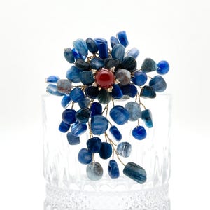 May include: A distinctive brooch with a central red gemstone, encircled by clusters of blue and grey stones. The stones are linked by fine gold-coloured wires, forming a floral or starburst pattern. The brooch is displayed against a clear glass background.