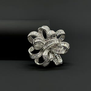 Può includere: Una spilla argentata a forma di fiocco, incastonata con numerose gemme chiare e scintillanti. Il design del fiocco presenta più anelli e pieghe, creando un effetto tridimensionale. La spilla è posta su uno sfondo scuro.