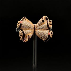 Puede incluir: Un broche de lazo dorado con diamantes de imitación transparentes y burdeos. El lazo está en un soporte transparente sobre un fondo negro.