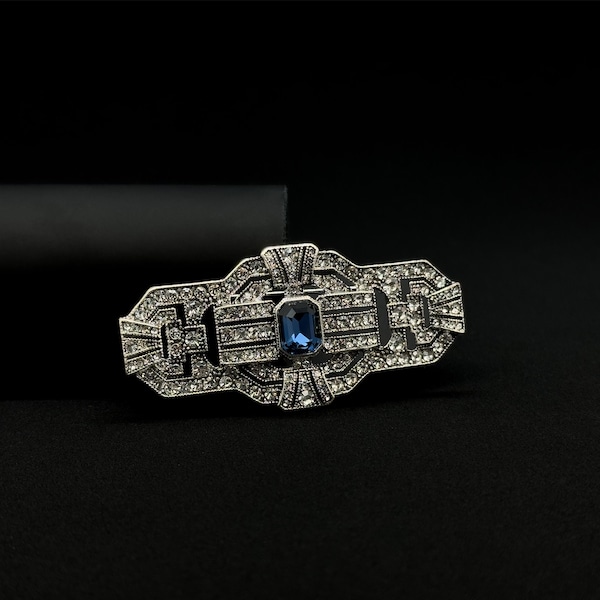 May include: A silver-toned Art Deco brooch with a rectangular blue gemstone center. The brooch is encrusted with small, clear crystals and features geometric designs. The background is black.