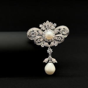 May include: A silver-toned brooch featuring a central round white pearl surrounded by clear, sparkling crystals in a symmetrical, ornate wing-like design. A single, larger teardrop-shaped white pearl hangs from the bottom, suspended by a crystal.