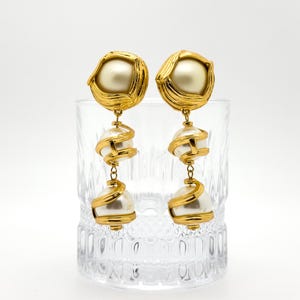 May include: A pair of gold-toned earrings with a layered design. Each earring features a large, pearlescent bead at the top and bottom, connected by a spiraling gold accent. The earrings are displayed in a clear glass.