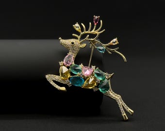Könnte beinhalten: Goldfarbene Hirschbrosche in Laufpose, besetzt mit klaren und farbenfrohen Edelsteinen. Die Brosche hat ein detailliertes Design mit einer Nadel auf der Rückseite für sicheren Halt. Die Edelsteine sind in den Farbtönen Rosa, Blau, Gelb und Grün gehalten.