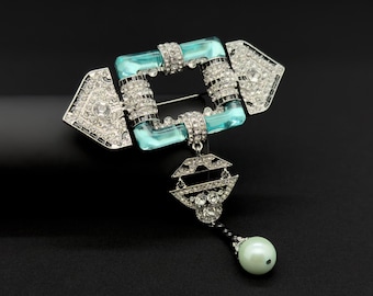 May include: An Art Deco style brooch with a square, light blue center and sparkling rhinestone accents. The brooch features a dangling geometric pendant and a faux pearl drop. The metal is silver-toned.