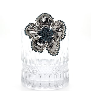 Pode incluir: Um broche de flor de prata com strass azuis. O broche está sobre uma superfície de vidro transparente.