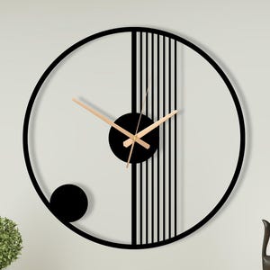 Peut inclure: Une horloge murale circulaire dotée d'un cadre en métal noir avec un cercle central noir et des lignes verticales. L'horloge possède de fines aiguilles des heures, des minutes et des secondes de couleur or rose. Un petit cercle noir est situé en bas.
