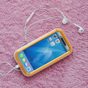 May include: A smartphone with a blue screen and a yellow and white case rests on a pink, fluffy surface. White earbuds and a charging cable are connected to the phone. The screen displays app icons.