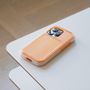 Può includere: Una custodia per telefono arancione e bianca con un design a coste sul retro poggia su un tavolo bianco. La custodia ha un design protettivo attorno alle lenti della fotocamera. Il tavolo è appoggiato su un pavimento in legno chiaro.