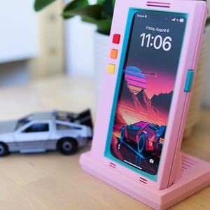 May include: Pink phone stand with a retro design, featuring a phone displaying a colorful car graphic. The stand has red, orange, and yellow buttons. A silver toy car is in the background. The phone screen shows "Friday, August 8, 11:06".
