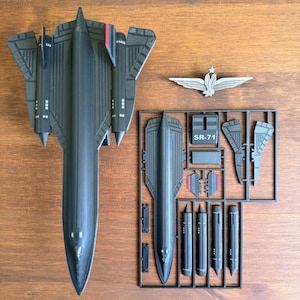 Puede incluir: Un modelo de avión SR-71 negro con detalles rojos, un emblema plateado y piezas sin montar en un marco de inyección. El modelo se muestra sobre una superficie de madera. El texto "SR-71" es visible en el marco.