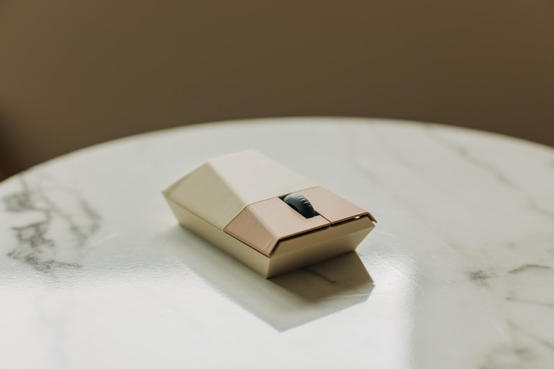 Puede incluir: Un rat&oacute;n inal&aacute;mbrico para ordenador de color blanco y beige con un dise&ntilde;o geom&eacute;trico redondeado. El rat&oacute;n tiene un solo bot&oacute;n y una rueda de desplazamiento.