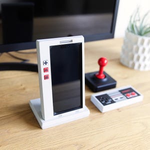 Może przedstawiać: Biały stojak na smartfona z czerwonymi i czarnymi przyciskami, przypominający klasyczny kontroler do gier wideo. Czerwony joystick i mały kontroler również znajdują się na drewnianej powierzchni. Stojak trzyma czarny ekran.