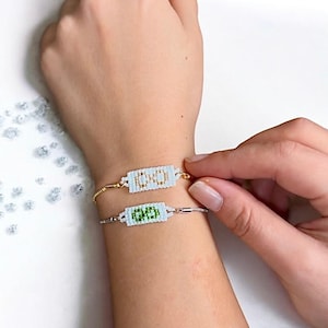 May include: Two beaded bracelets with white beads and gold accents. The top bracelet has the word "BOOM" in gold beads. The bottom bracelet has the word "LOVE" in green beads.