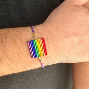 May include: A beaded bracelet featuring a rainbow flag design. The bracelet has a rectangular centerpiece with red, orange, yellow, green, blue, and purple beads. The bracelet is attached to a purple cord with gold-colored metal accents.