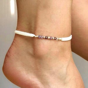 May include: A white macrame anklet with small purple and silver beads. The anklet is worn on the ankle. The macrame is a thick, woven design. The beads are small and round.