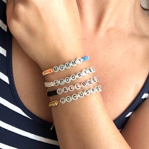 Pode incluir: Quatro pulseiras artesanais com contas de letras brancas que soletram "MUMMY BY", "SPOOKY BY", "HALLOWEE" e "HOCUS POCUS". As pulseiras estão em um pulso, com cordão colorido e detalhes dourados. A pessoa está usando uma regata listrada azul e branca.