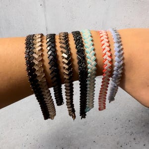 Op de afbeelding: Een set van acht kralenarmbanden in verschillende kleuren, waaronder zwart, wit, bruin, rood en blauw. De armbanden zijn gemaakt van kleine, vierkante kralen die in een visgraatpatroon zijn geweven.