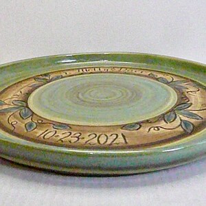 May include: A handmade ceramic plate with a light green rim and a central design. The center is a light blue-green with concentric circles. The plate features a brown band with leaf and vine designs and the date "10-23-2021".