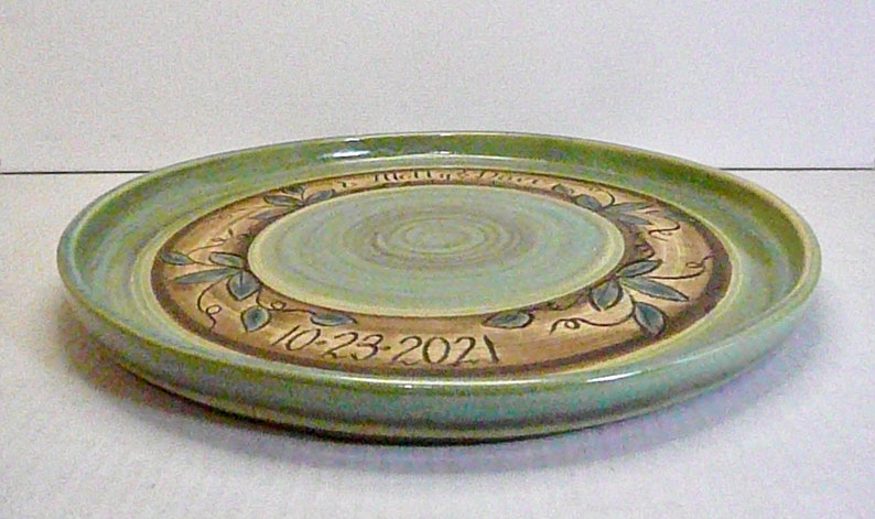 May include: A handmade ceramic plate with a light green rim and a central design. The center is a light blue-green with concentric circles. The plate features a brown band with leaf and vine designs and the date "10-23-2021".