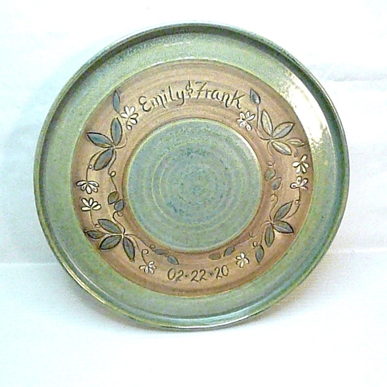 May include: A handmade ceramic plate with a green and tan color scheme. The plate features a floral design around the center, with the names "Emily & Frank" and the date "02-22-20" inscribed.