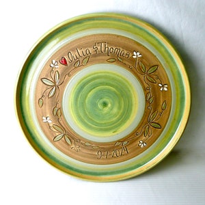 May include: A handcrafted ceramic plate with a green and brown spiral design. The plate features the names "Julia & Thomas" and the date "9-7-2024", along with a floral wreath design and a red heart.