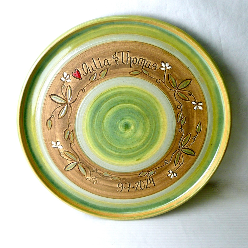 May include: A handcrafted ceramic plate with a green and brown spiral design. The plate features the names "Julia & Thomas" and the date "9-7-2024", along with a floral wreath design and a red heart.