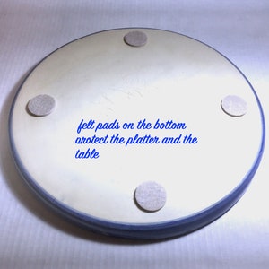 May include: A round, off-white ceramic platter with a blue rim. The bottom of the platter has four gray felt pads. Text in blue reads "felt pads on the bottom protect the platter and the table."