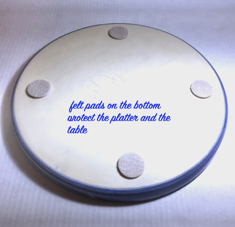 May include: A round, off-white ceramic platter with a blue rim. The bottom of the platter has four gray felt pads. Text in blue reads "felt pads on the bottom protect the platter and the table."
