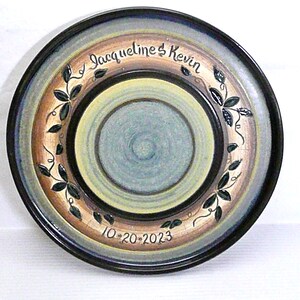 May include: A decorative ceramic plate with a hand-painted design. The plate features concentric circles in shades of blue, tan, and yellow, with a black rim. The text "Jacqueline & Kevin" and the date "10-20-2023" are inscribed on the plate.