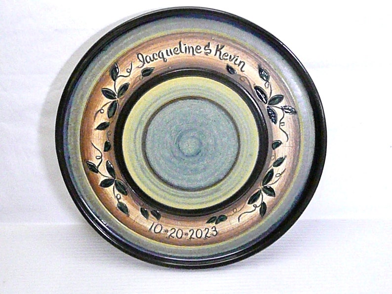 May include: A decorative ceramic plate with a hand-painted design. The plate features concentric circles in shades of blue, tan, and yellow, with a black rim. The text "Jacqueline & Kevin" and the date "10-20-2023" are inscribed on the plate.