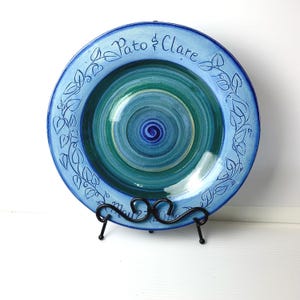 May include: A round ceramic plate featuring a blue and green swirled center and a wide blue rim decorated with hand-painted vine motifs. The text Pato & Clare and May 27 2026 are inscribed on the rim. The plate rests on a black metal stand.