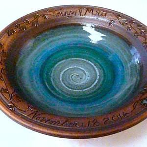 May include: A handmade ceramic bowl with a brown rim and a blue and green interior. The bowl features a spiral design in the center and is inscribed with "Casey & Matt" and "November 18 2016" around the edge. Decorative leaf patterns are carved around the rim.