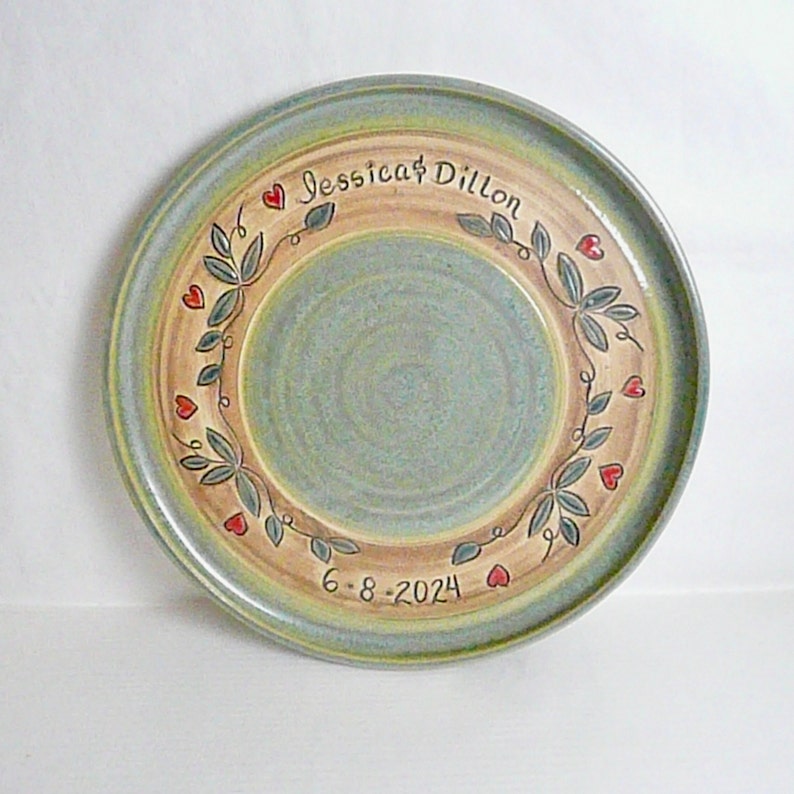 May include: A handcrafted ceramic plate with a light blue and tan color scheme. The plate features a decorative wreath of leaves and hearts, with the names "Jessica & Dillon" and the date "6-8-2024" inscribed.