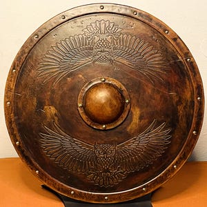 May include: A round, wooden shield with a detailed owl design. The shield is a dark brown color with a central boss and brass rivets around the edge. The owl design features outstretched wings.