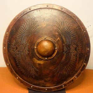 May include: A round, antique-style shield with a dark brown finish. The shield features an embossed design of two symmetrical eagle wings. A central, circular boss and brass rivets complete the design. This item is suitable for display or costume.